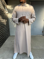  Homme portant un qamis saoudien couleur sable à manches longues, col officier boutonné et poche poitrine, pour un style raffiné et moderne.