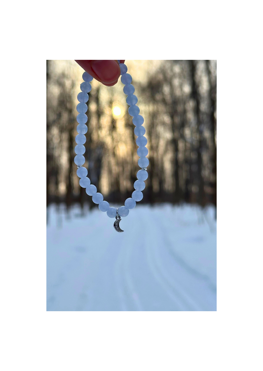 Bracelet tasbih en perles bleu givré avec pendentif lune argentée, suspendu dans un paysage enneigé au coucher du soleil
