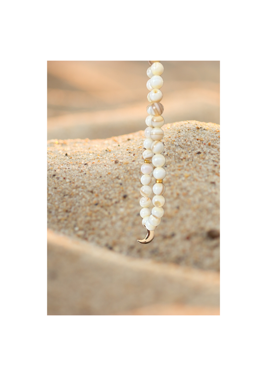 Bracelet tasbih en perles nacrées blanches avec pendentif lune dorée, suspendu au-dessus de sable doré