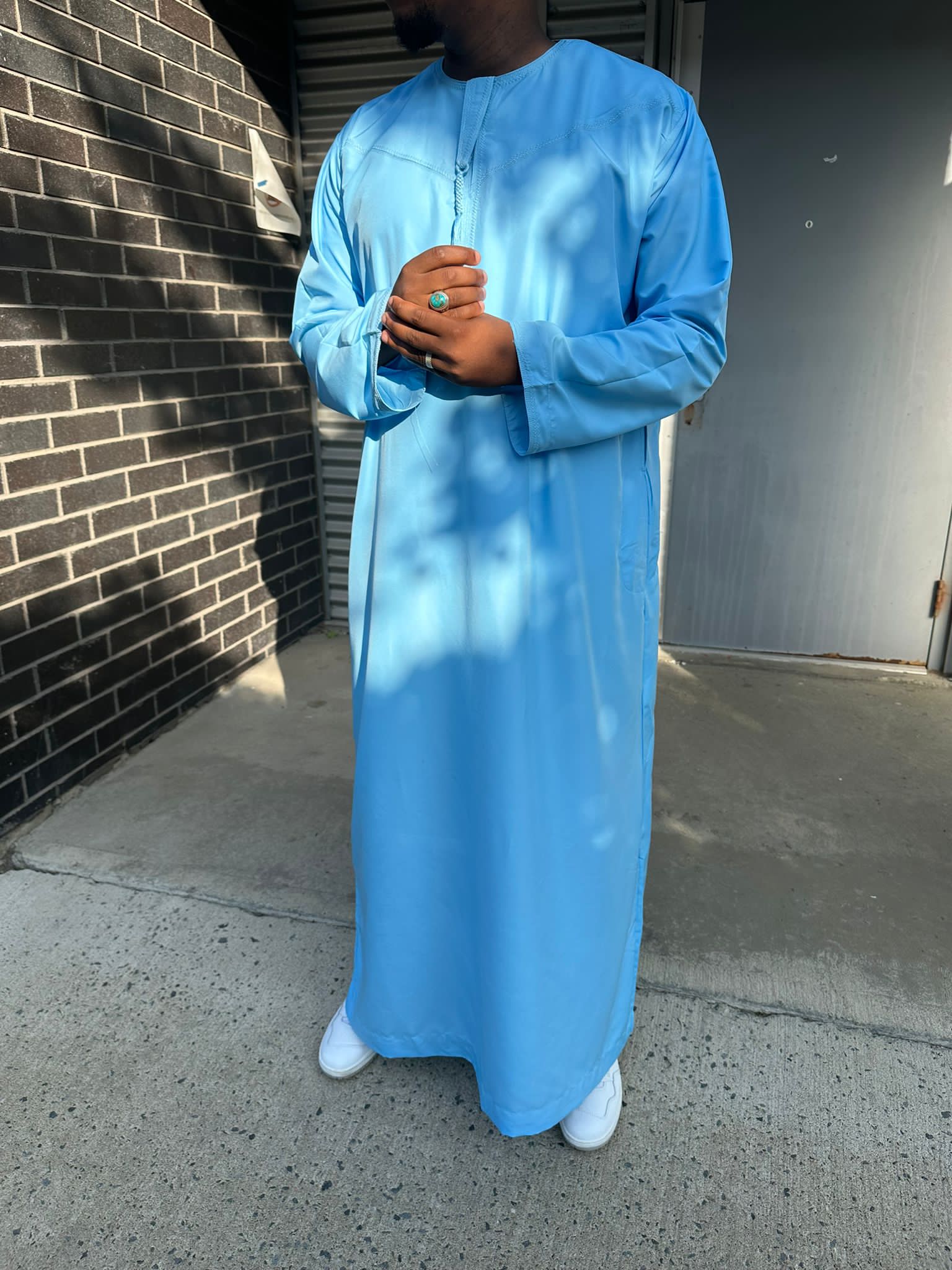  Homme portant un qamis bleu ciel traditionnel, debout avec les mains jointes, en extérieur.