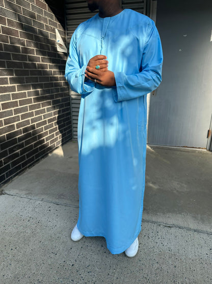  Homme portant un qamis bleu ciel traditionnel, debout avec les mains jointes, en extérieur.