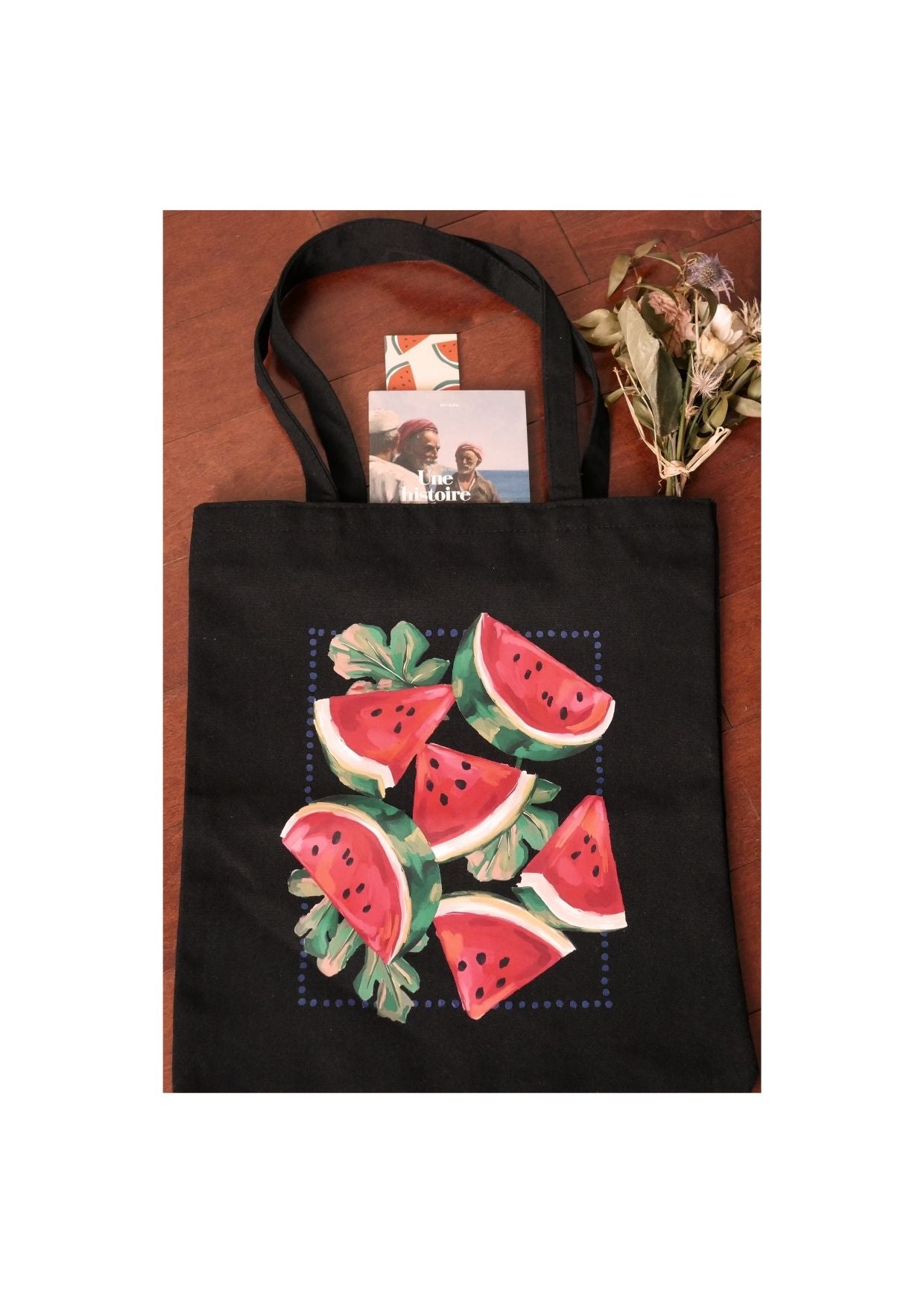 Sac tote en toile noire avec imprimé de tranches de pastèque, accompagné de fleurs séchées et de livres sur une surface en bois.