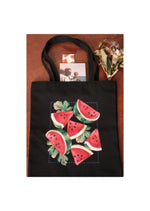 Sac tote en toile noire avec imprimé de tranches de pastèque, accompagné de fleurs séchées et de livres sur une surface en bois.